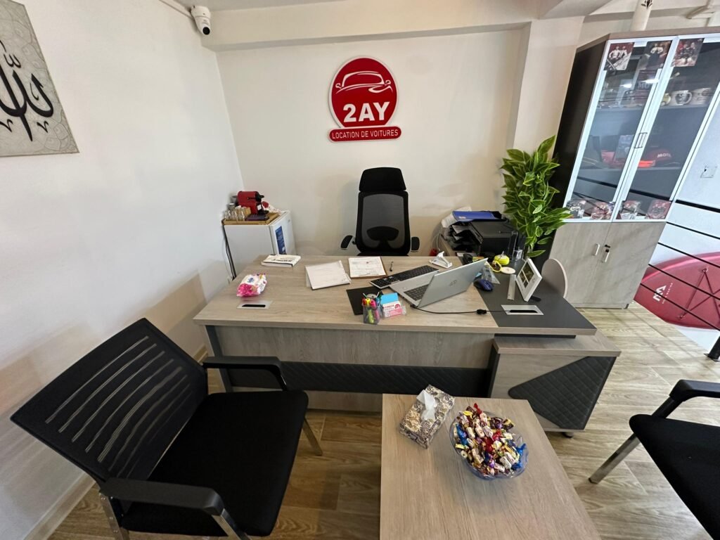Poste de travail de l'agent 2AY Location De Voitures à Sétif — laptop, contrats de location, logo 2AY visible au mur, agence professionnelle de location de voiture en Algérie
